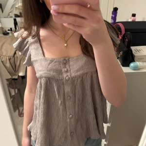 Grå blus med volangärmar - Säljer en söt grå blus med volangärmar och knappar framtill. Blusen har en luftig passform och är perfekt för varmare dagar. Den har en lätt rynkad textur som ger en avslappnad look.