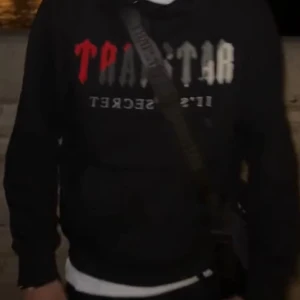 Svart hoodie från Trapstar - Hjälper min kille att sälja sin svarta hoodie från Trapstar med texten 'It's a Secret' i vitt och rött på framsidan. Köptes på Plick för ca 1000, endast en defekt vilket är att plasten på det ena snöret är sönder av förra ägaren, den går att klippas av om det stör😊(se bild 4) annars är den i topp skick, hör av er vid funderingar