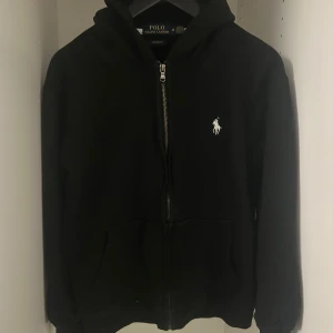 Svart hoodie från Ralph Lauren - Säljer en stilren svart hoodie från Ralph Lauren med dragkedja och det klassiska vita logotypen på bröstet. Perfekt för en avslappnad look. Den har långa ärmar och en praktisk huva. Det är M men sitter väldigt litet så skulle säga <S
