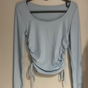 Ljusblå ribbad topp - Säljer en ljusblå ribbad topp med långa ärmar och knytband i sidorna för en justerbar passform. Perfekt för en stilren och bekväm look.