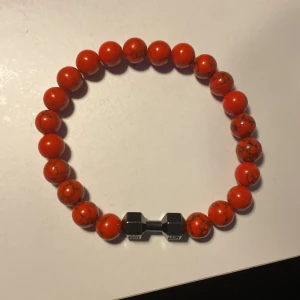 Rött pärlarmband med hantel - Snyggt armband med röda pärlor och en liten hantel i metall som detalj. Perfekt för att ge en sportig touch till din stil.