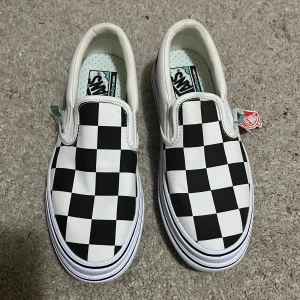 Svartvita slip-on sneakers från Vans - Snygga slip-on sneakers från Vans med klassiskt svartvitt rutmönster. Skorna har en bekväm ComfyCush-sula och är perfekta för en avslappnad stil. Den ikoniska designen gör dem till ett måste i garderoben. Helt nya oanvända med etikett på