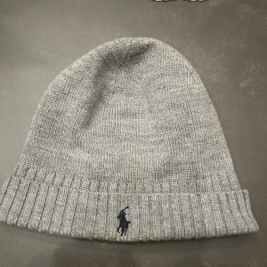 Grå stickad mössa från Polo Ralph Lauren - Snygg grå stickad mössa från Polo Ralph Lauren med broderad logga. Perfekt för kyliga dagar med sin ribbade kant som ger en skön passform. En stilren accessoar för både höst och vinter.