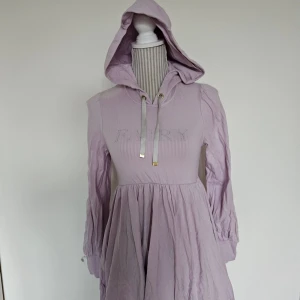 Lila klänning med huva från E Hyphen World Gallery - Klänning från E Hyphen World Gallery med långa ärmar och huva. Står ''FAIRY'' på den. Begagnat men bra skick förutom att den blekts lite på två ställen (se bild), spår av användning finns men hel och ren. ''Onesize'' men uppskattar att den passar XXS-M beroende på önskad passform.  Rökfritt hem men har katter.