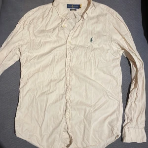 Beige skjorta från Ralph Lauren - Säljer en stilren beige skjorta från Ralph Lauren i slim fit. Skjortan har långa ärmar och är broderad med den klassiska loggan i grönt på bröstet. Perfekt för en elegant look.