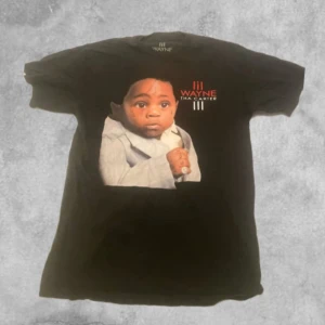 Svart t-shirt med tryck från Lil Wayne - (XS men väldigt stor i storleken så typ en medium) Cool svart t-shirt med ett tryck av ett barn i kostym och texten 'Tha Carter III' från Lil Wayne. Perfekt för fans av artisten. T-shirten har en loose fit och är kortärmad.