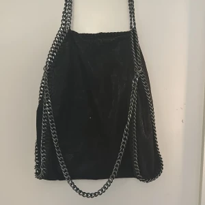 Svart axelväska med kedjedetaljer - Snygg svart axelväska med coola kedjedetaljer. Väskan har ett leopardmönstrat foder och en praktisk dragkedja inuti. Perfekt för att ge din outfit en edgy touch.