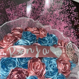 Bukett med konstgjorda rosor - En vacker bukett med konstgjorda rosor i rosa och blått, dekorerad med glitter. Rosorna är arrangerade i en genomskinlig plastförpackning med rosa blommönster. Ett rosa band med texten 'Ronja' och ett glittrigt hjärta pryder buketten.