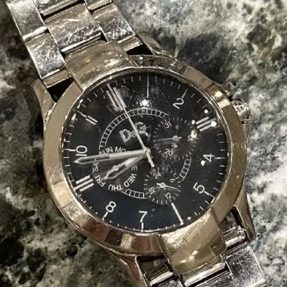 Snygg silverfärgad klocka från D&G med svart urtavla och flera små urtavlor för extra funktioner. Armbandet är i metall och har en klassisk design. Perfekt för den som vill ha en stilren accessoar. Klockan är gammal men går fortfarande och använda, finns defekter (smått vattenskadad därav priset) (repor) . Asusteet.