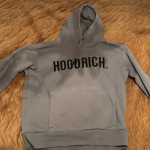 Grå hoodie från Hoodrich - Säljer en stilren grå hoodie från Hoodrich med svart text på framsidan. Den har en klassisk känguruficka och justerbar huva med dragsko. Perfekt för en avslappnad stil.