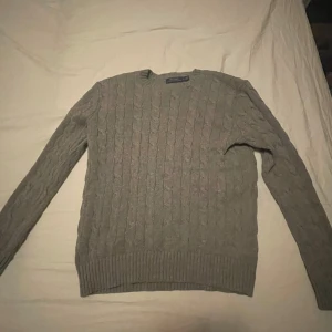 Grå stickad tröja från Polo Ralph Lauren - Säljer en stilren grå stickad tröja från Polo Ralph Lauren med klassiskt flätmönster. Tröjan har långa ärmar och en diskret broderad logga på bröstet. Perfekt för kyligare dagar.