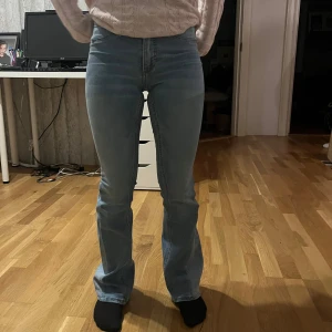 Blå bootcut jeans med rosetter - Snygga ljusblå bootcut jeans med hög midja och söta rosa rosetter på bakfickorna. Perfekta för en avslappnad stil med en twist. De har en klassisk en dragkedja med knapp framtill.