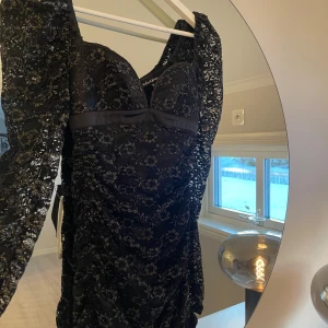 Svart spetsklänning från For Love & Lemons - Elegant svart klänning i spets från For Love & Lemons. Klänningen har en glittrig finish och en vacker blommig design. Den är figurnära med långa ärmar och en stilren siluett. Perfekt för en glamorös kväll!