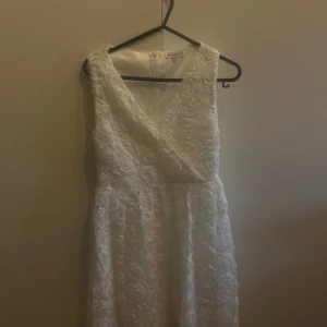 Vit student-klänning med handsydda blommor  - Elegant vit klänning med ett vackert blommigt mönster i tyget. Klänningen har en v-ringad design och är ärmlös, vilket ger en stilren look. Perfekt för studenten tillfällen. - Inga fläckar, i nyskick