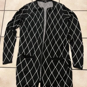 Svart och vit långärmad kofta - Snygg svart kofta med vita rutor och långa ärmar. Den har en öppen design utan knappar eller dragkedja och två fickor framtill. Perfekt för att ge en stilren touch till din outfit.