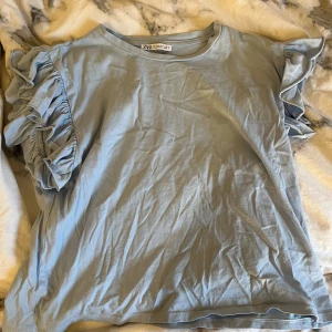 Blå T-shirt med volanger - Säljer min ljusblå T-shirt med volanger från Zara då den inte kommer till användning. Passar perfekt till alla möjliga kläders plagg.