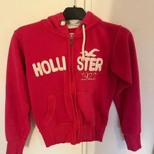 Rosa zip-hoodie från Hollister - Säljer en snygg rosa zip-hoodie från Hollister med dragkedja och vit logga framtill. Den har en mysig huva med grått foder och praktiska fickor. Perfekt för en avslappnad stil.