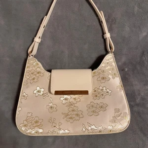 Beige axelväska med blommigt mönster - Elegant beige axelväska med justerbar rem och vackert blommigt mönster i guld. Väskan har en stilren design med en praktisk ficka på insidan och ett rymligt och rent inre. 