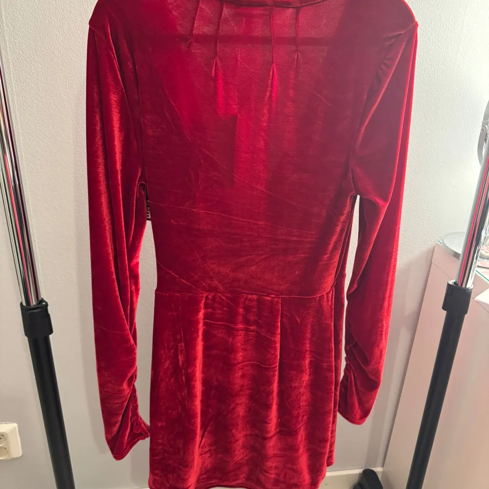 Snygg röd omlottklänning i sammet från Nelly. Klänningen har långa ärmar och en v-ringad design med knytband i midjan. Perfekt för att sticka ut med sin djupa färg och mjuka material. Aldrig använd och prislappen sitter kvar!. Mekot.