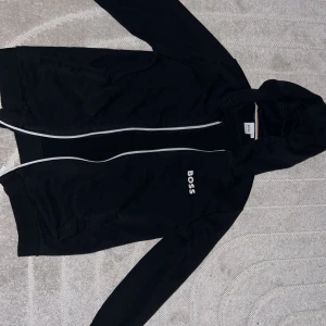 Svart hoodie från Boss - Säljer en stilren svart hoodie från Boss med dragkedja och huva. Jackan har långa ärmar och en vit logotyp på bröstet. Perfekt för en avslappnad look.