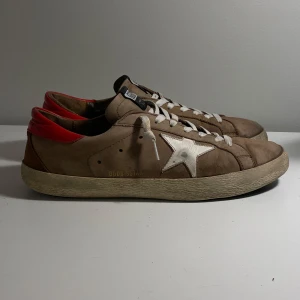 Golden Goose - Tjena! Säljer nu mina beige sneakers från Golden Goose med vit stjärndetalj på sidan och röd häl. Skorna har vita snören och en klassisk design med en sliten look. Perfekta för en avslappnad stil.
