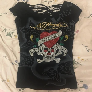 Svart Ed Hardy topp med dödskallemotiv - Snygg svart topp från Ed Hardy med ett stort dödskallemotiv och rosor. Toppen har ett unikt korsat banddesign på ryggen och är kortärmad. Perfekt för en edgy stil!