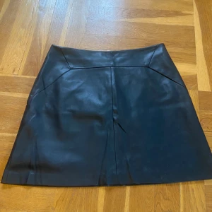 Svart kjol från Lindex - Snygg svart kjol från Lindex i skinnimitation. Kjolen har en stilren design med dragkedja i baksidan och en diskret söm på framsidan. (Hög i midjan)