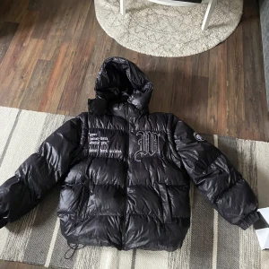 Svart dunjacka med tryck - Säljer en svart dunjacka med huva och tryck på bröstet och ryggen. Jackan har en pufferdesign med långa ärmar och dragsko i nederkant för justerbar passform. Man kan dra av armarna å använda den som väst. Storlek S sitter perfekt, skick 10/10 använd 1 gång bara. Priset är inte hugget i sten