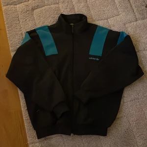 Adidas zip up - Adidas tröja i bra skick, inga defekter! Grå med turkosa ränder vid axlarna. Skriv för fler bilder eller frågor. (Tveka inte på att prisförhandla!!)
