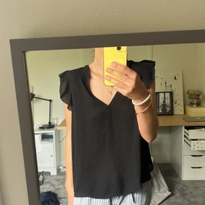 Svart blus  - En svart blus från H&M, den har volanger vid axlarna och är V-ringad. Den är svart då den ser lite grå aktig ut på bilden, priset kan diskuteras vid snabb affär!