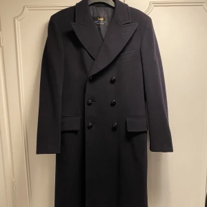 Junex rock i ull - Rejäl, mörkblå, herr rock med dubbelknäppning från det svenska märket Junex. Räcker ner under knäet. Rocken är kemtvättad.  Uppskattas vara i storlek medium. 