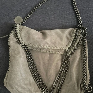 Stella McCartney Falabella Fold Over Tote  - Äkta Stella McCartney väska i fin gråbrun färg ”Shaggy deer” med gunmetal-färgade kedjor. Tillhörande plånbok medföljer. Den ena knappen som sluter väskan är skadad - se bild 3. I övrigt i väldigt gott skick. 