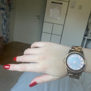 Guldig Marc Jacobs klocka  - I bra skick,med rosa och guldiga stenar på ordet”marc”. Justerbar samt tjockt guldig armband runt. Inte bytt batteri på länge så kan behövas