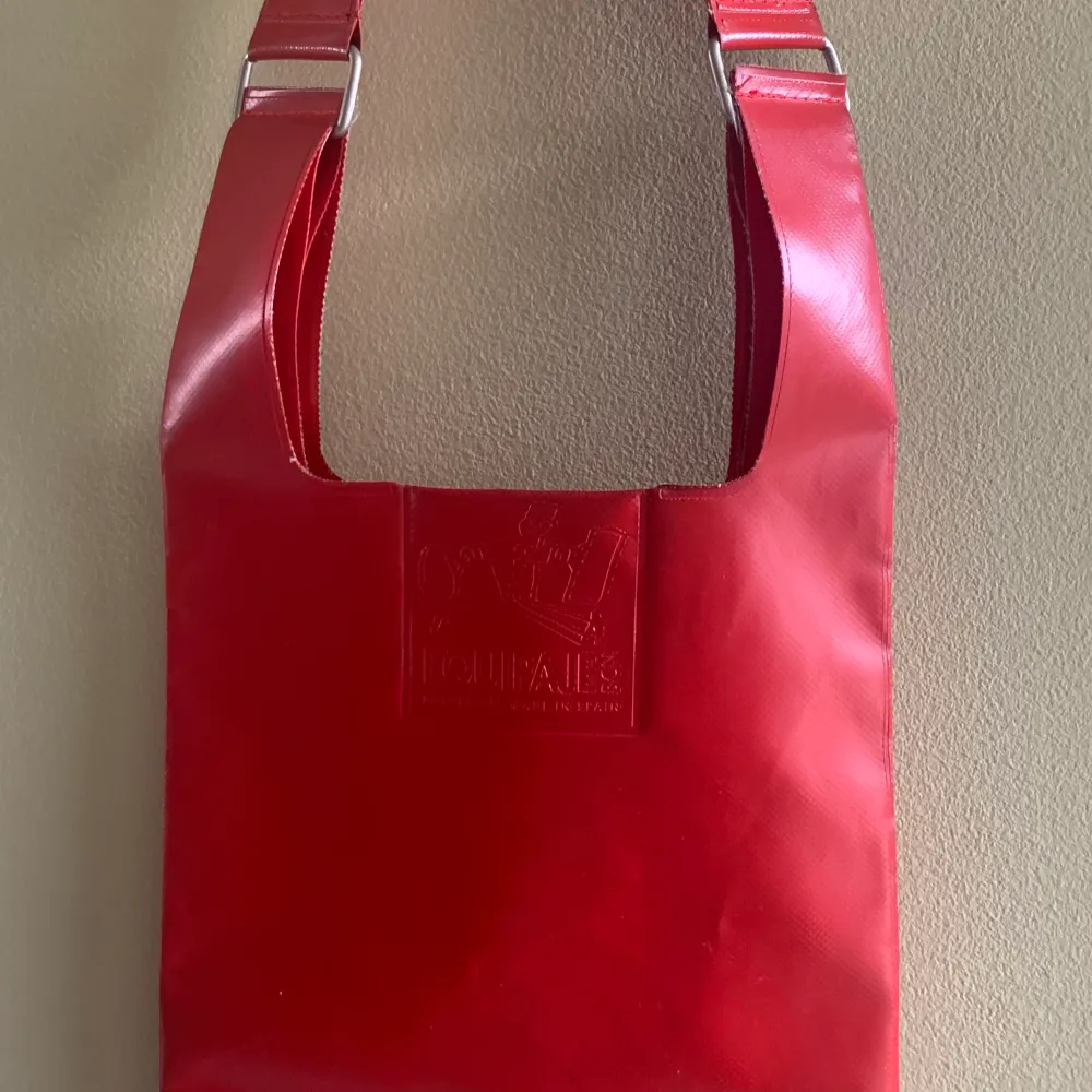 Röd axelväska i plast från Equipaje BCN. Knappt använd, men har fått ett litet märke på axelremmen, se bild. 35x24 cm är själva väskan, förutom remmen.. Väskor.