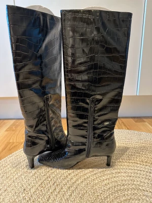 NA-KD Boots med vida skaft  - Knälånga boots med låg klack ifrån NA-KD. Har ett bredare knähögt skaft, en halv dragkedja på insidan och en krokodil effekt.  Oanvända Orginalpris: 815 kr