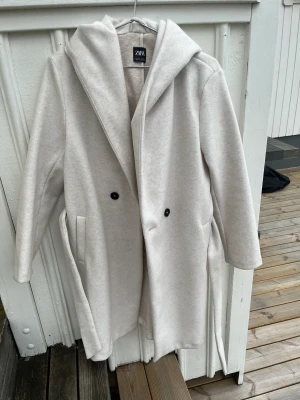 Beige Zara kappa - Ursnygg lång kappa ifrån zara i en snygg kräm vit färg. Storlek S men passar även XS och M. Säljes då den inte passat min stil och därför inte kommit till användning. Endast använd 2 gånger. Alltså fortfarande i nyskick.  