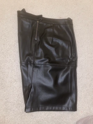 Kjol i PU-läder - Snygg kjol i svart PU-läder som slutar strax ovanför knäna. Liten slit baktill. Fina dekorativa sömmar framtill. Använd fåtal gånger!