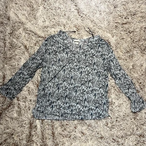 Vit/blå blus med blå fågelmönster H&M - Supersöt vit/blå blus från H&M med blåa fåglar och svarta grenar som mönster. Blusen har v-ringning med små knappar framtill och långa ärmar med lätt volang vid ärmslut. Tillverkad i tunn, luftig polyester – perfekt för en chill och stilren look. Tröjans material är väldigt luftigt och lätt och även stretchig. Storlek M