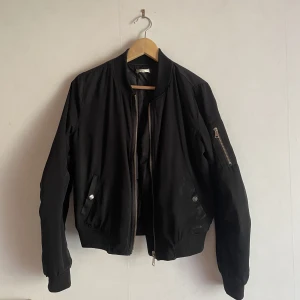 Svart klassisk bomberjacka - Klassisk svart bomberjacka med cool guldig dragkedja i fram. Fodrad med silke/satin vilket gör den väldigt skön att ha på sig. Perfekt för en sensommarkväll eller när hösten börjar smyga sig på. 