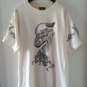 Vintage t-shirt med drake - Vintage vit t-shirt med drake och tribaler i storlek L. Den är lite gulnad och har några fläckar (se bild 2), går nog troligen bort lite i tvätten. Fraktkostnad blir 47kr.