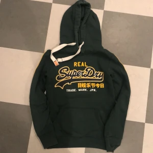 Superdry hoodie - Mörkgrön hoodie i fint, använt skick. Lite ”sprickor” i trycket. Köparen står för frakten. 