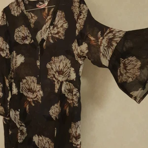 Mörkgrön tunika i chiffong - Tunika i från Indiska i chiffong. Aldrig använd med prislapp kvar. Ordinarie pris 499 kr, 3/4 ärmar med volanger, runkor i sidan av midjan, knäppning endast upptill, går till knäna på mig som är 163 cm. Kan bäras till byxor eller klänning med något under då den är lite genomskinlig. Bakgrunden är mörkgrön, gröna blad, vita stora blommor med några orangea inslag. Xs men oversized (bättre på någon i stl. S).