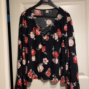 Blommig blus strl 42 - Jättefin svartblommig blus från H&M med justerbar snörning över bysten. Vida armar. Aldrig använd. Köparen betalar för frakt