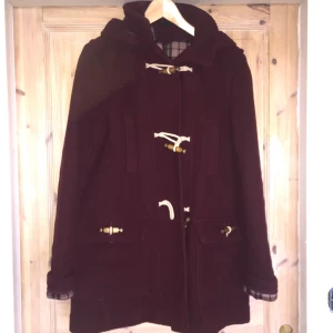  - Vinröd duffelkappa i 80% ull, gott skick och väldigt varm. Kapuschongen kan man ta av. Två stora fickor. Knappar och duffelknappar. Fraktas med Skicka lätt för 75kr spårbart. Frakt tillkommer. Eller mötes upp i Stockholm. 
