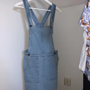 Jeansklänning i snickarbyxbodell - Ljusblå hängselklänning perfekt för sommaren. Finns inte längre att köpa. Använd ett fåtal gånger men nu för liten för mig tyvärr. Köparen står för frakt :)