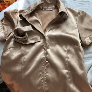Guldig blus   - Guldig blus i storlek 40. Köpt på second hand, men jag har endast använt den 1 gång! Följer med ett band att knyta i midjan!☺️ Saknar en knapp näst längst ner. 25kr 