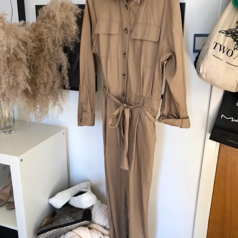 En beige jumpsuit med långa ben och armar från H&M, tillhörande snöre att knyta i midjan och bruna knappar upptil, även dragkedjan nedtill under knapparna för bättre passform! I väldigt bra skick! Passar mig som har storlek S/M och är 170 cm lång.. Övrigt.