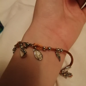 Armband med smycken - Fint, litet armband i glansig guldbrun tråd med hängen i silver med buddhistiska drag🧡💛 frakt ingår