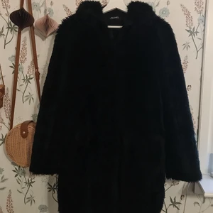 Svart kappa i fuskpäls, Monki - Välanvänd pälsjacka från Monki med huva och knäppning. Storlek S. Mått: Bredd 100 cm, längd 89 cm. 