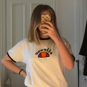 Ellesse t-shirt  - Ellesse tröja i str 38 med en broderad halvmåne i orange+röd. Den har även svarta detaljer i kragen och mudden på ärmarna. 